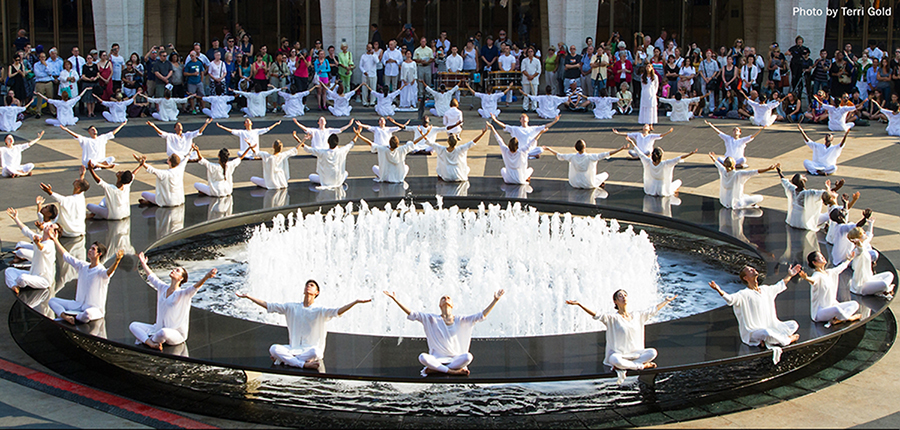 A group of people dressed in all white sit around a fountain in a circle with legs crossed and arms raised in the air. Two more circles of people in the same clothing and position surround them, and an audience stands around at the back.